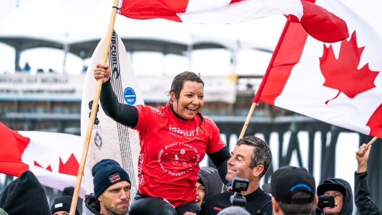 A woman wearing a wetsuit smiles while holding up a Canadian flag, with two men holding her up on their shoulders.