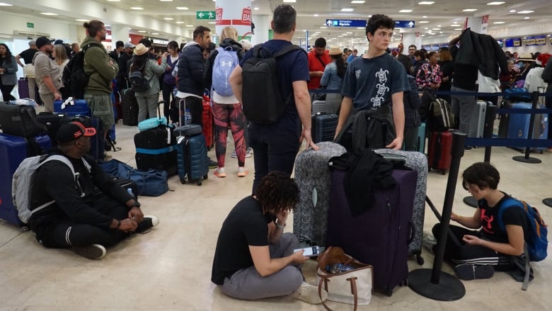 A lot of people sit or stand around luggage in an airport. Some people are sitting on the floor looking at their cellphones.