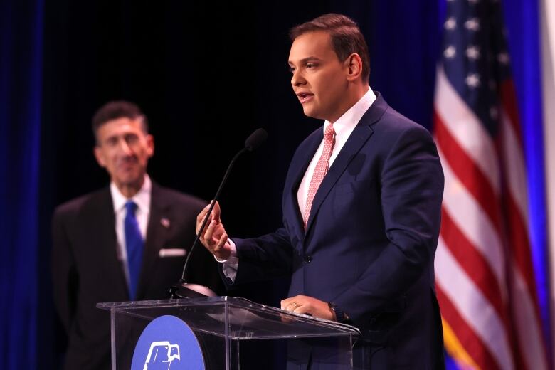 A man in a dark blue suit and red tie speaks into a microphone at a lectern while another man looks on.