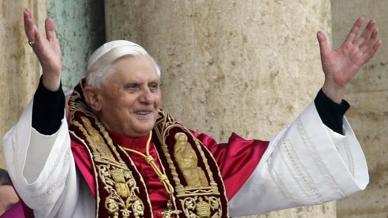 White-haired man wearing white clergy robe with gold and red scarves over top stands with arms outstretched. 