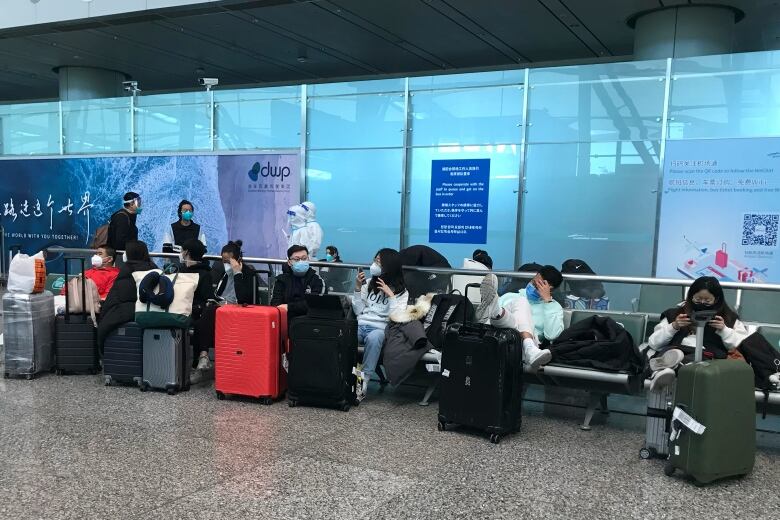 People wearing masks sit in a row of seats with suitcases. 