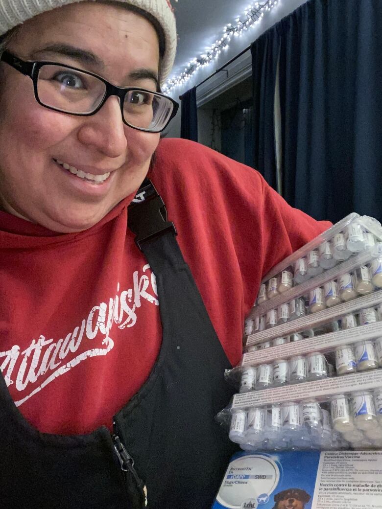 Phoebe wears a red sweatshirt and snow pants with suspenders and holds a case of parvovirus vaccines wrapped in plastic. She smiles widely.