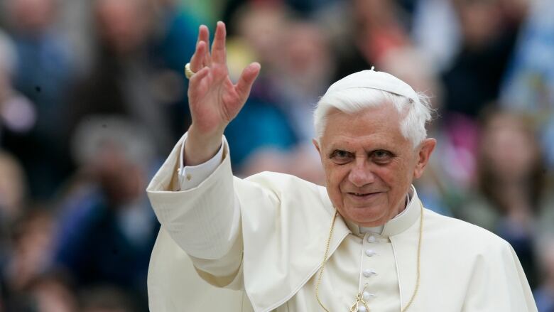 Pope Benedict XVI, dressed in cream-coloured robes and a hat and a gold cross around his neck, raises his right arm.