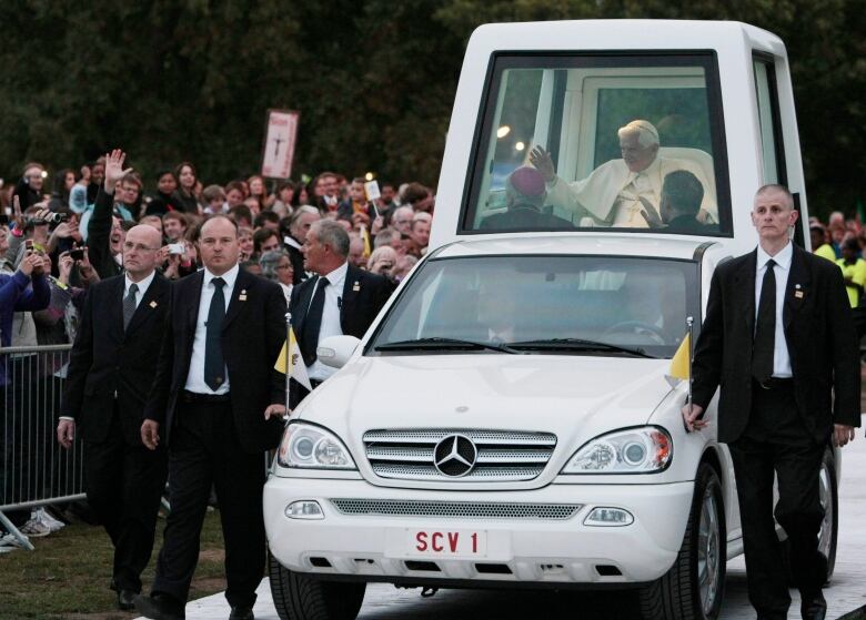 A man in cream robes sits in a white tower attached to the back of a white Mercedes vehicle. The car is surrounded by bodyguards wearing black suits. Members of the public can be seen waving signs behind barricades.