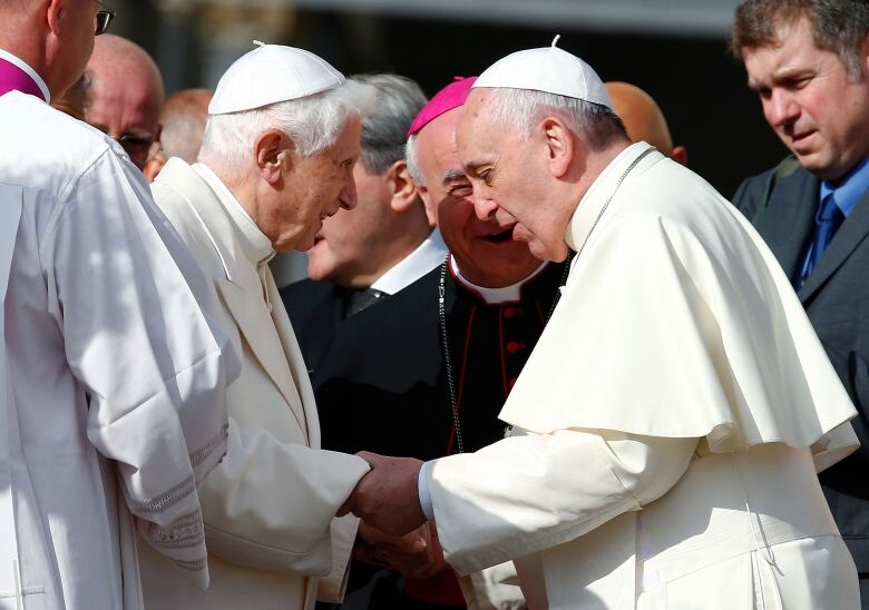 Two elderly men in cream-coloured outfits face each other, holding hands, while others look on.