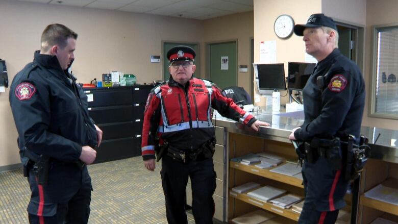 Three police officers stand next to a counter while the man in the middle appears to be speaking.