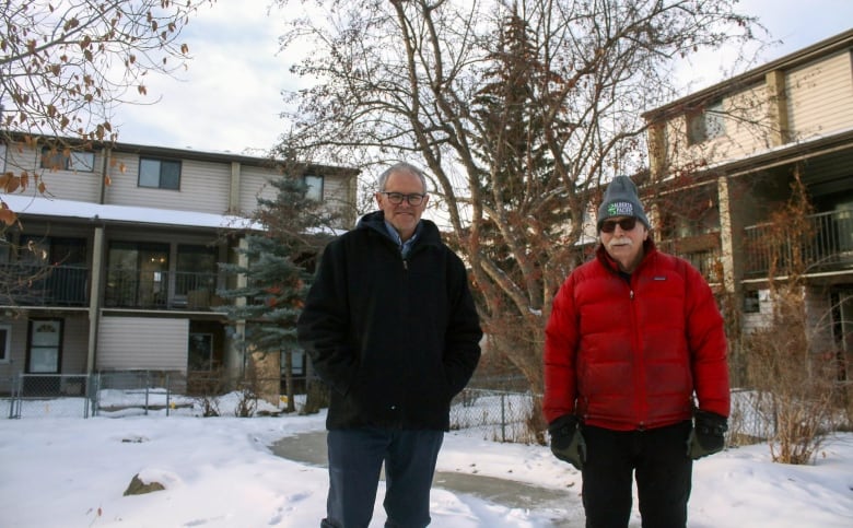 Two men stand outside, in front of brown townhouses, in the winter. One is wearing a black jacket, the other a red jacket with a toque and gloves.