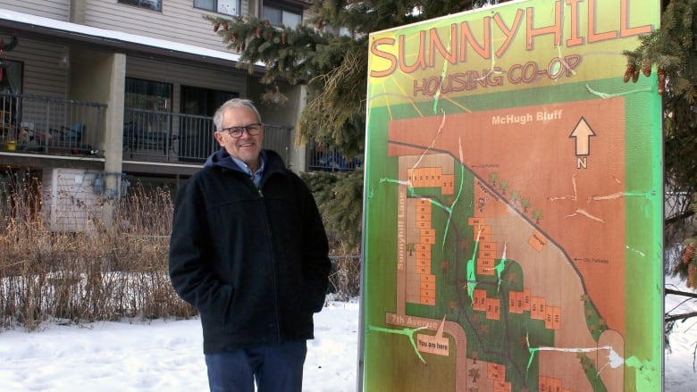 A man stands outside, smiling, next to a large green and orange sign that reads, Sunnyhill Housing Co-op.