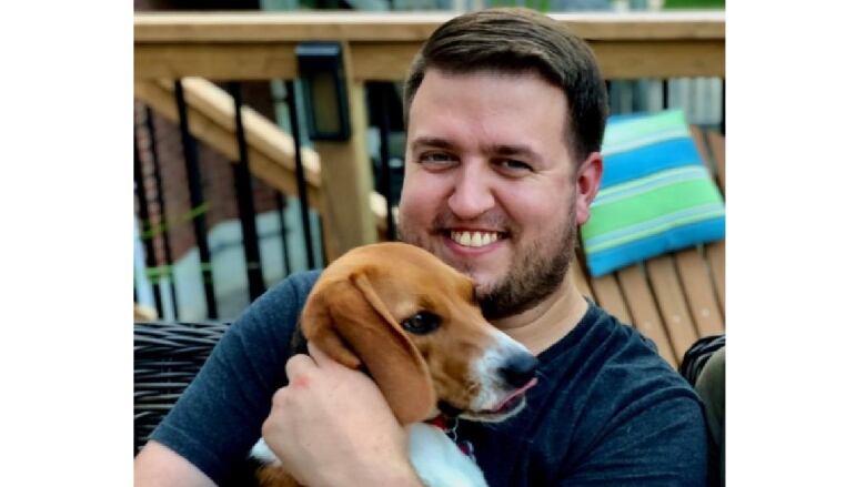 A man with dark hair and a dark beard smiles while looking at the camera. He's holding a beagle.