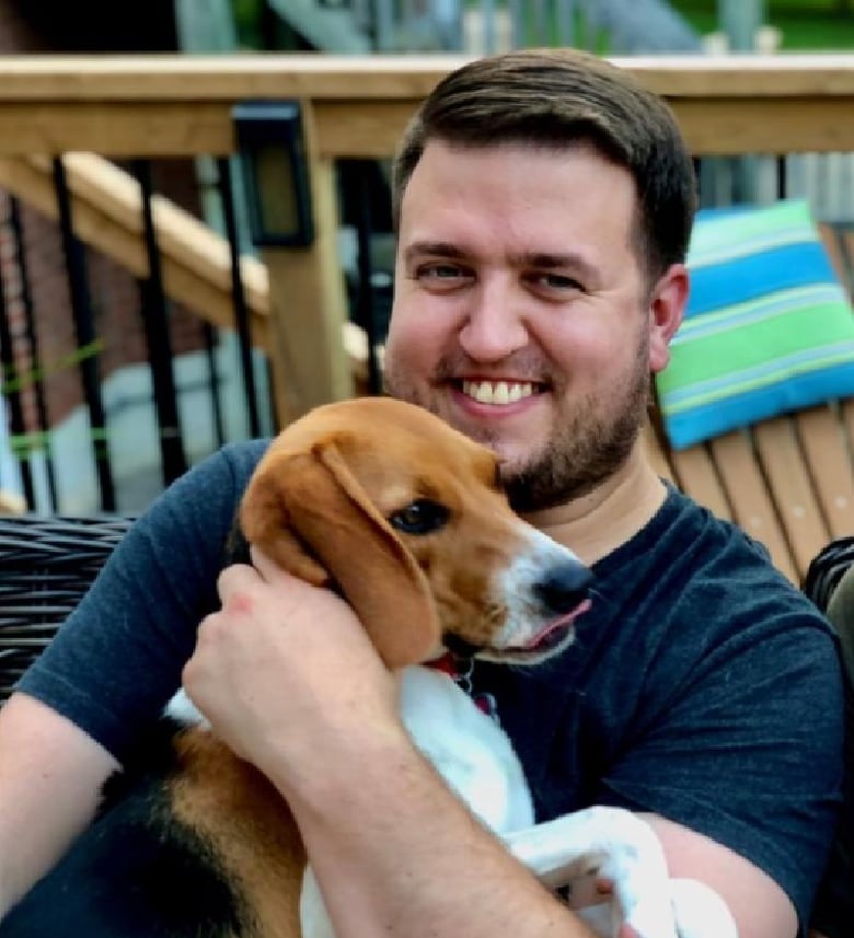 A man with dark hair and a dark beard smiles while looking at the camera. He's holding a beagle.