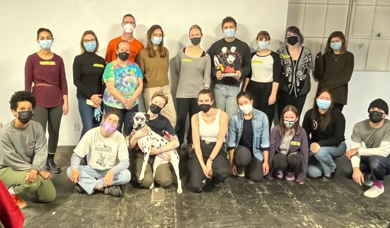 A group of people pose against a flat white wall. Everyone is wearing masks, with one person in the front holding a Dalmation. 