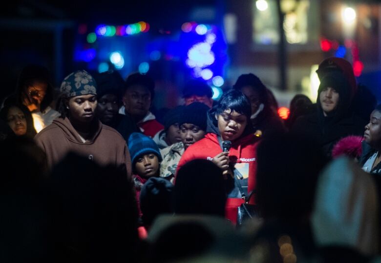 A crowd of people stand in vigil for Nicous D'Andre. A woman, looking down, holds a microphone.