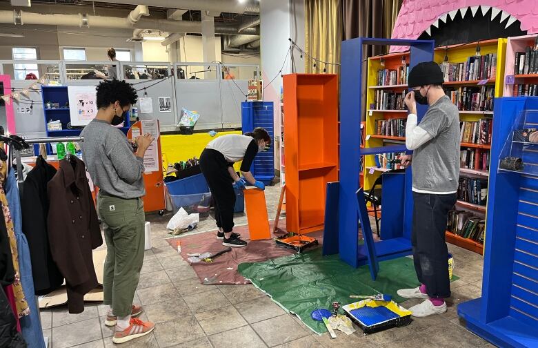 Three people are working away at painting bookcases. On the left, a person with curly cropped hair wearing a mask scrolls through their phone. In the centre, a person bends over to paint a bright orange shelf. On the right, a person adjusts his mask. 