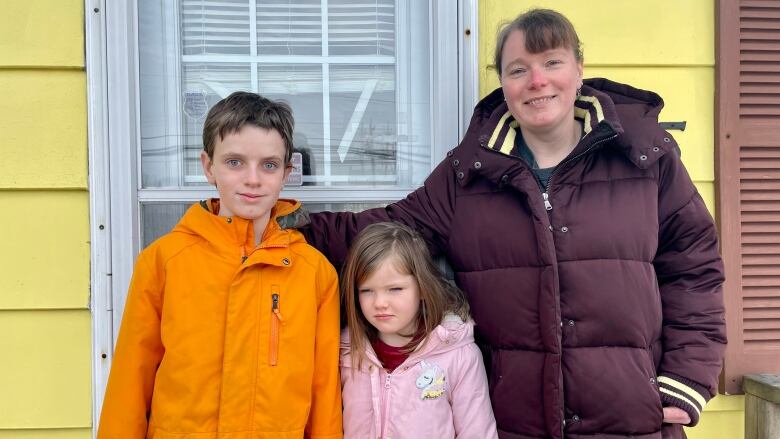 A mother and her two young children are seen smiling in front of their yellow home.