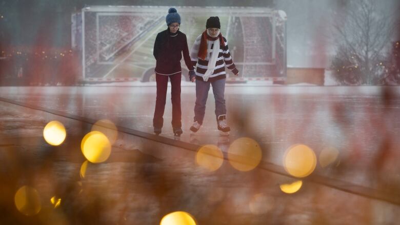 Two people hold hands skating on an outdoor rink on a misty day. There are holiday lights in the foreground.