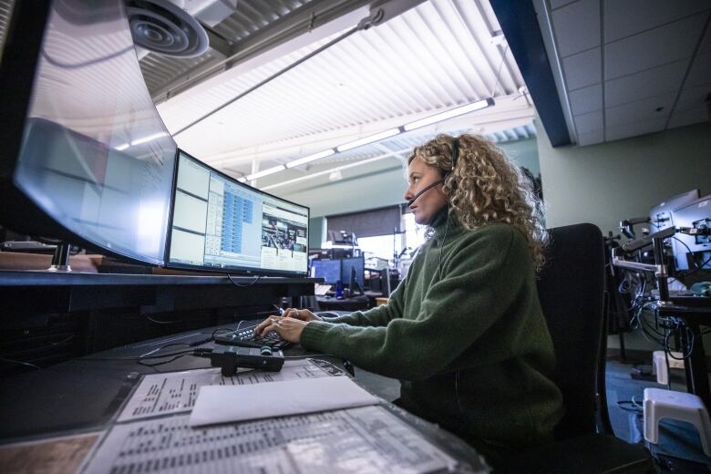 A woman sits in front of a computer with a headset and several monitors in front of her.