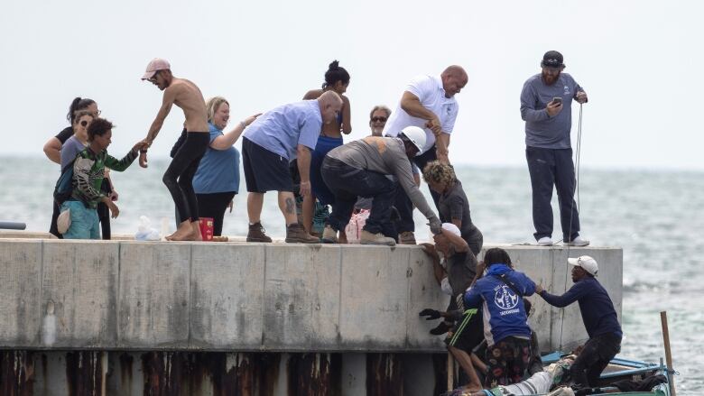 Several people on a dock reach down to lift people on a small vessel in the water.