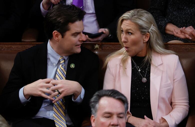 A dark haired man with a blue and yellow striped tie speaks to a surprised looking woman with long blond hair, wearing a pink jacket. 