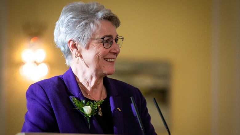 A woman with white hair in a blue blazer stands at a podium and smiles.