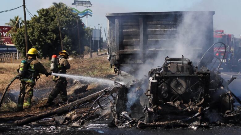 A firefighter points a fire hose at a burned-out vehicle as another firefighter walks in front of them.