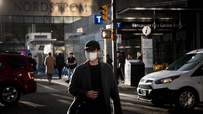 A man wearing a black hat and glasses crosses the street in downtown Vancouver while wearing a white respiratory mask. 