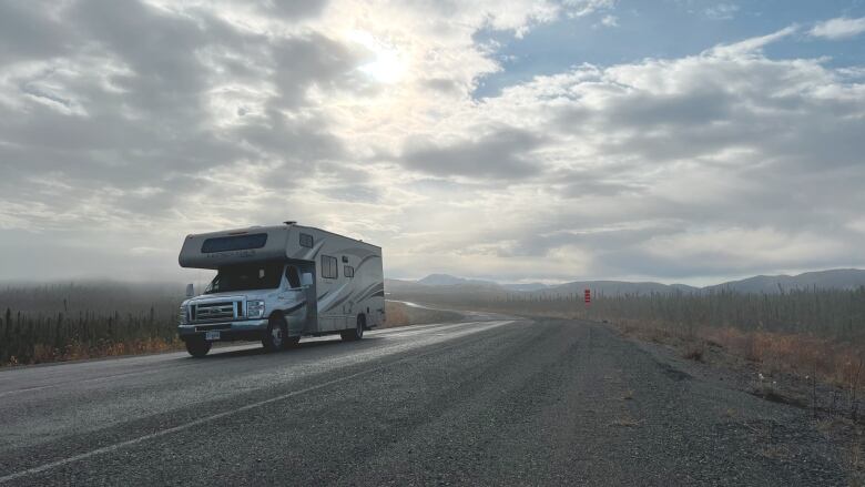 An RV drives on an empty road with mountains and trees in the background.