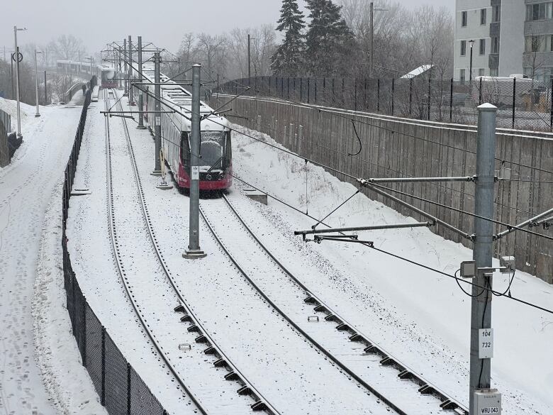 Two stopped light rail trains on a track between stations. All are covered by a dusting of snow.