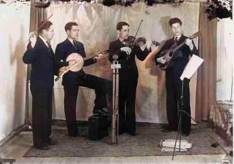 An old photo showing four men in suits standing in front of a single microphone, with a music stand off to one side. One man plays a banjo, one is playing a violin and the third is playing a guitar. The fourth man holds a wind instrument in his left hand and is conducting the other three with a baton in his right hand.