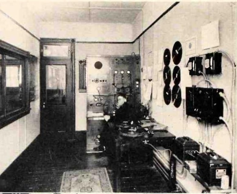 An old photo showing a long room, with a large wall to wall transmitter at the far end. An engineer sits in a chair next to a table, where a turntable sits. Large batteries and various electronic equipment sit in the foreground.