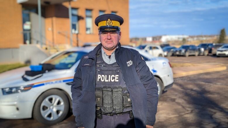 A police officer stands in front of his vehicle, wearing a uniform vest and hat and posing for the camera.