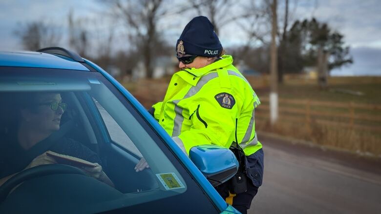 An RCMP officer in a bright yellow reflective jacket and sunglasses speaks with a person in a vehicle who has rolled down their window.