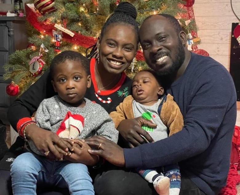 A man and a woman sitting on a red rug with two young boys on their laps. They are sitting in front of a lit-up Christmas tree.