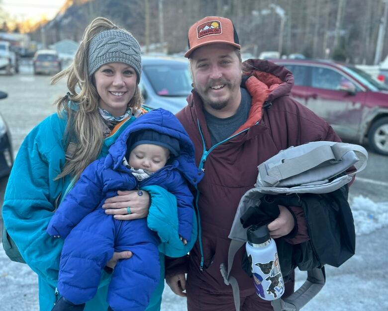 A man and woman holding a sleeping baby stand outside in winter coats.