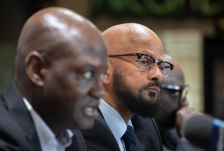 Three men sit next to each other, two men are blurred and the man in the middle is in focus.  