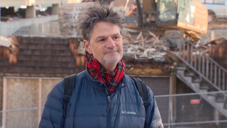 A man wearing a blue puffer jacket and a red tartan scarf stands in front of the demolition, he's looking to the right of the frame off-camera.