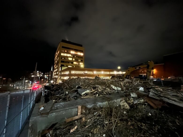 A nighttime picture of the site where a house has been demolished, showing just a pile of boards and city lights.