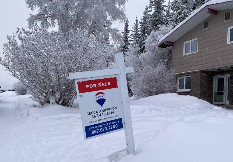 A for sale sign sits on a snowy lawn in front of a house.