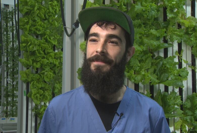 A man in scrubs speaks with a reporter in a warehouse filled with rows of hydroponically grown produce , such as lettuce and basil.