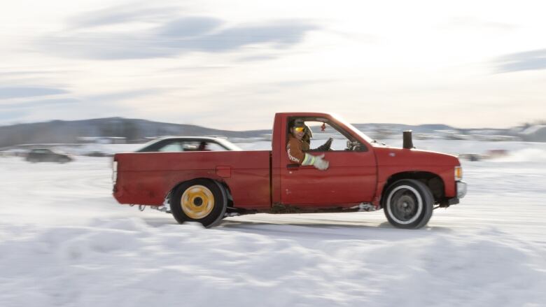 A red pickup truck is seen in profile driving along the ice. Inside are two people, one of which is wearing polarized reflective sunglasses. 