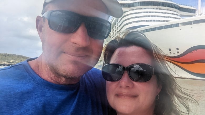 A man and woman, both wearing sunglasses, pose for the camera. A large cruise ship is in the background.