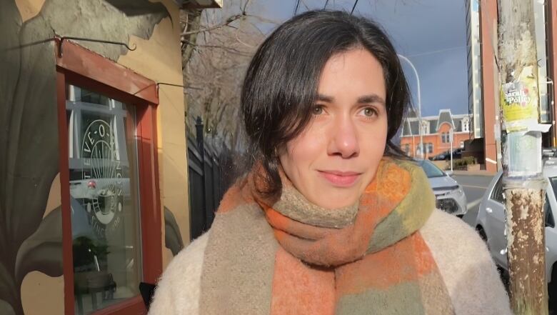 A woman with dark hair stands outside a cafe on a city street. She is wearing a beige coat and green and orange check scarf