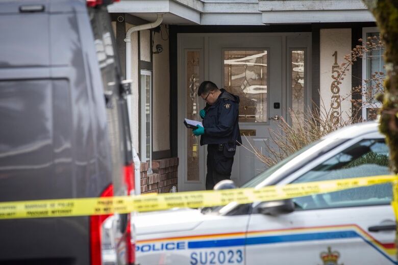 A police officer stand behind police tape at a house's front door and looks in his coat pocket.