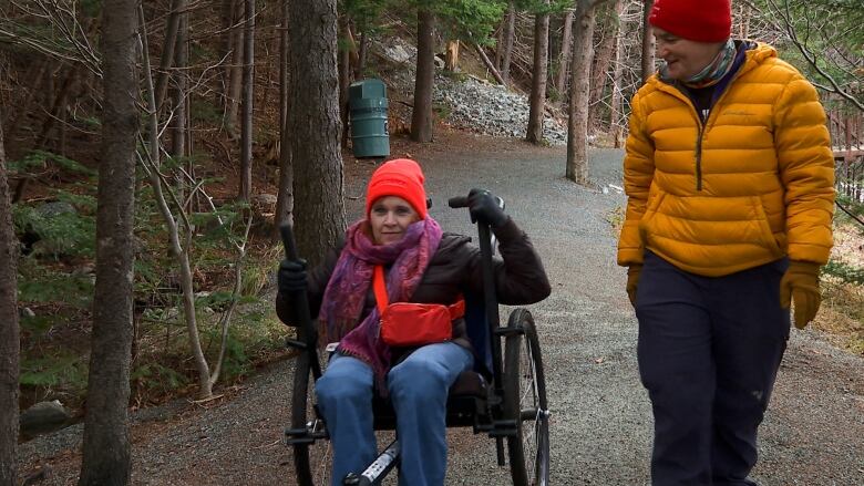 Two women on a hiking trail, one walking and one riding in a self-propelled wheelchair.