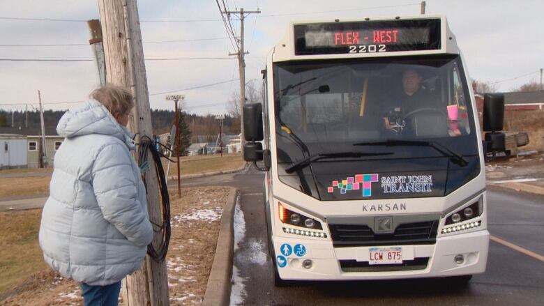 A bus pulls up to the curb while a women in a blue coat waits to get on. The bus has a Saint John Transit logo and lights that say FLEX west. 