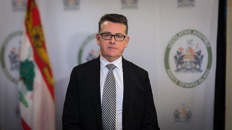 A man in a suit and tie stands in front of a backdrop with the P.E.I. Legislative Assembly logo and the P.E.I. flag.