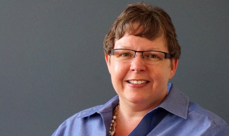 A woman wearing a blue shirt, glasses and multiple necklaces smiles before a grey backdrop. 