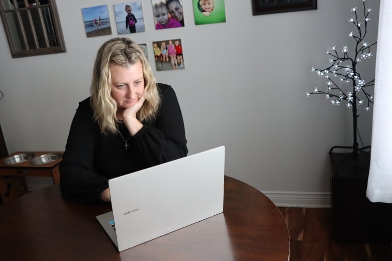 A blonde woman sits in front of a laptop at her kitchen table. 