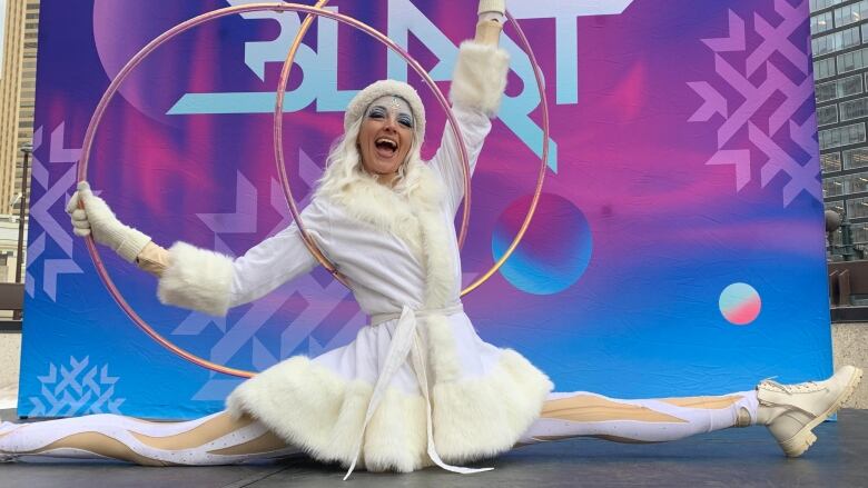 A woman dressed in white tights and a wintry white robe with fur-like cuffs performs the splits while holding two hula-hoops over her head. 