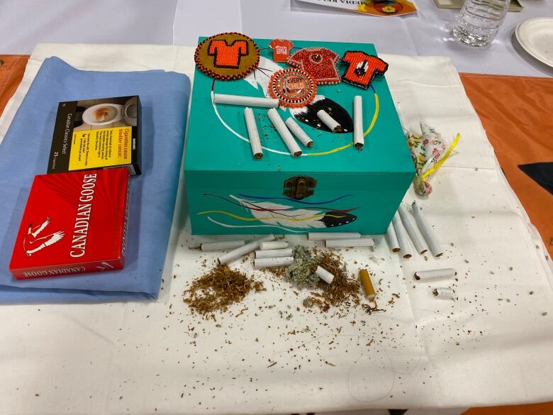 A turquoise box with a feather painted on it sits on a white-clothed table. To the left are two packs of cigarettes. There are cigarettes and tobacco laying beside the box and on top of it. Homemade ornaments showing orange t-shirts are also placed on top of the box.