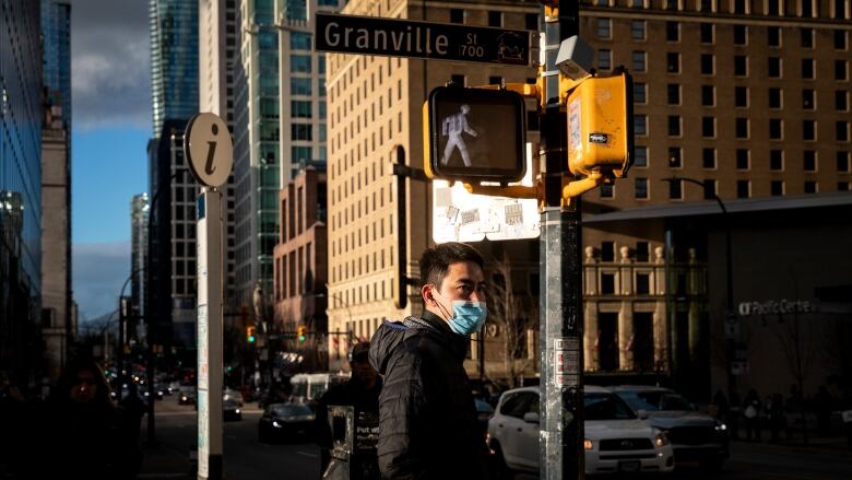 A man wearing a surgical mask stands at a crosswalk in downtown Vancouver.